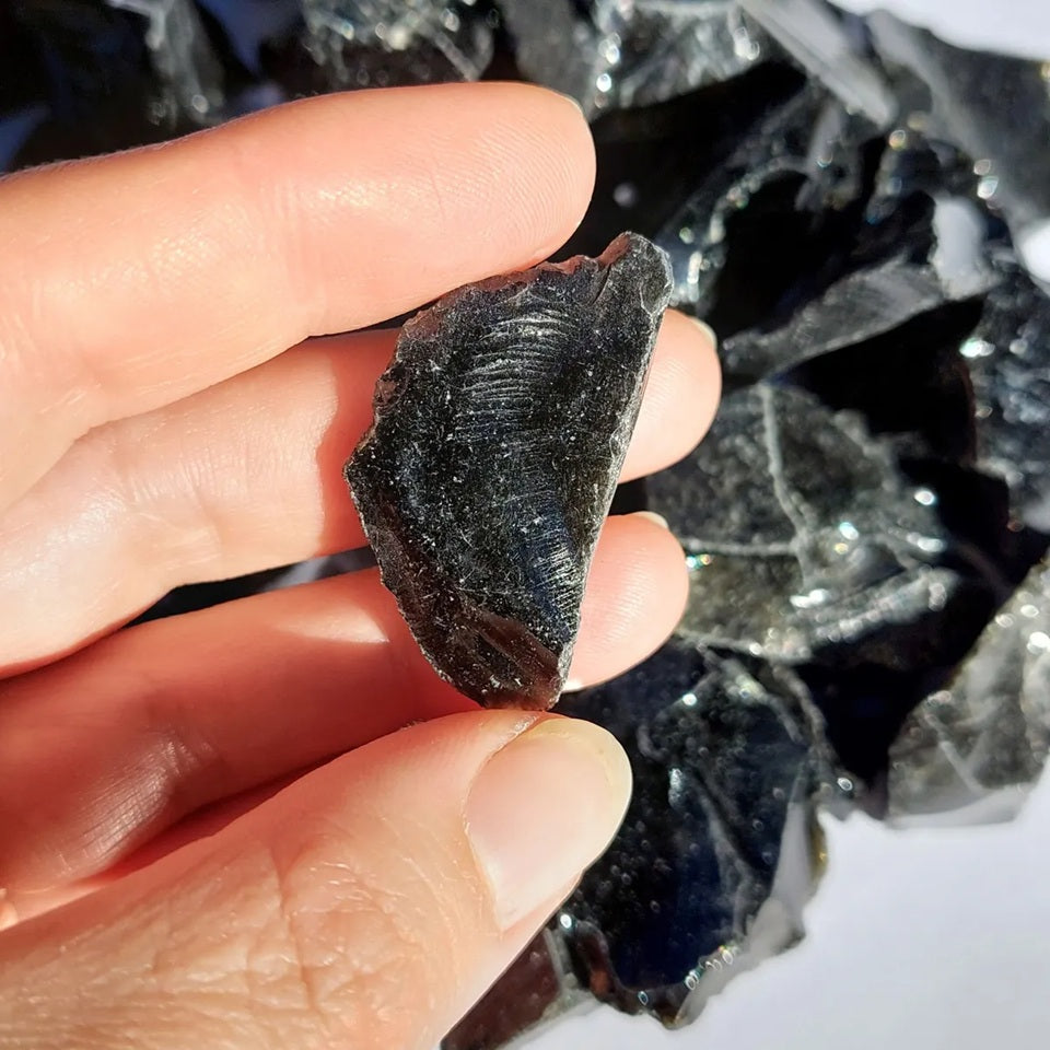 Pieza de obsidiana negra en bruto sostenida con los dedos, de forma plana e irregular con estrías finas y brillo vítreo
