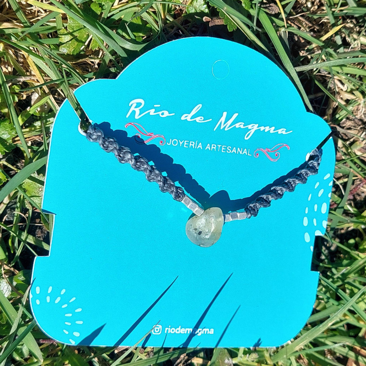 Collar Luz de prehnita natural en macramé gris con cuentas de plata 950, mostrando la piedra verde translúcida como elemento principal, sobre tarjeta de Río de Magma