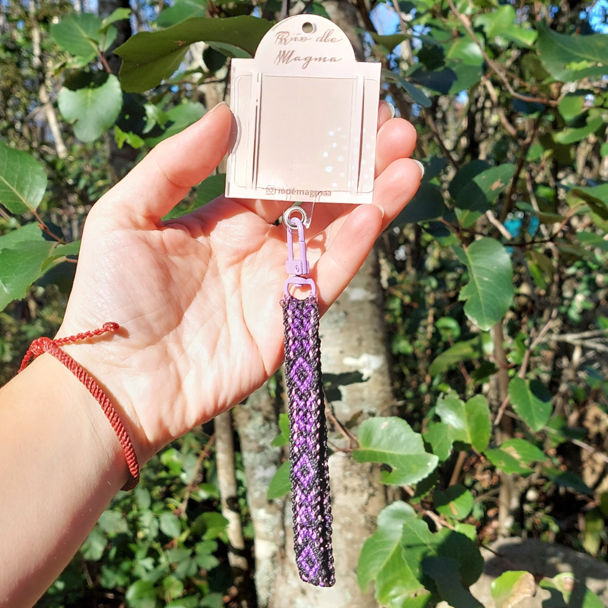 Celular con correa artesanal de macramé morado sostenido en la mano, fotografiado en exterior.