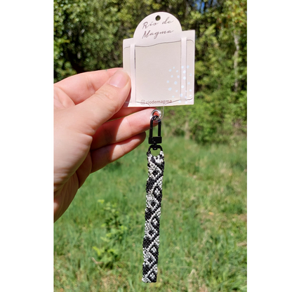 Adaptador para carcasa de celular con correa de macramé negro y plateado colgando, fotografiado al aire libre.
