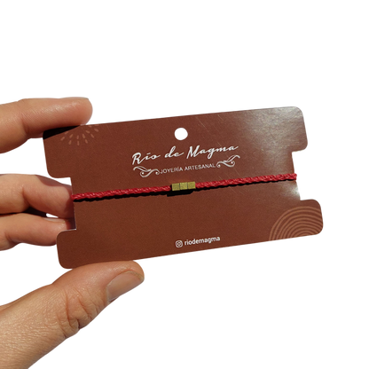 Pulsera minimalista con cuentas de bronce en hilo rojo, montada en tarjeta Río de Magma, fondo blanco